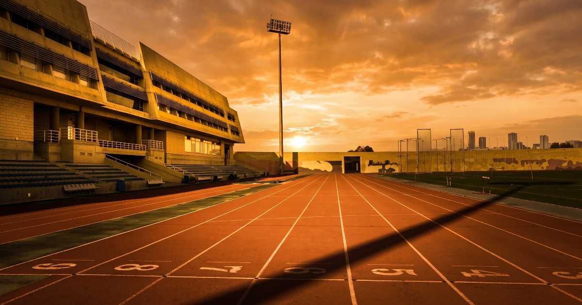 Empty running track at sunset with glowing orange sky, lane numbers visible in the foreground and stadium grandstand on the left